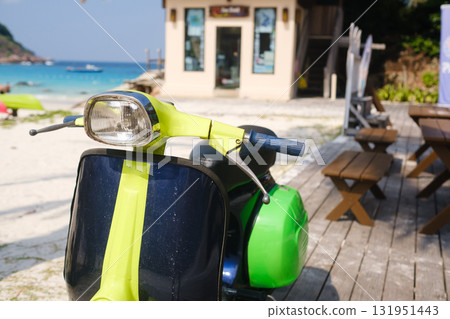 Bright scooter parked near a beachside cafe with clear blue water and wooden tables in the background during a sunny day Bright scooter parked near a beachside cafe with clear blue water and wooden tables in the background during a sunny day 131951443