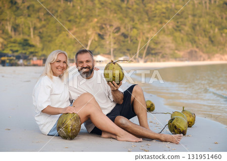 Couple enjoying a sunny beach day while relaxing and holding coconuts on the shore of a tropical paradise 131951460