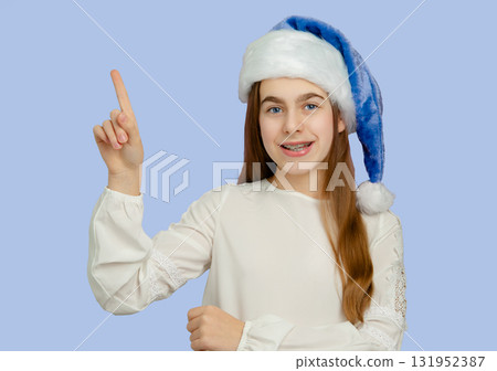 Smiling girl with blue Santa hat on blue background. Festive portrait for Christmas and winter celebrations. Smiling girl with blue Santa hat on blue background. Festive portrait for Christmas and winter celebrations. 131952387