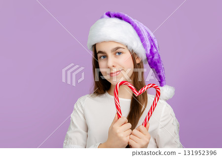 Festive girl in purple Santa hat holding Christmas sugar sticks candy in front of her eye on pastel background. 131952396