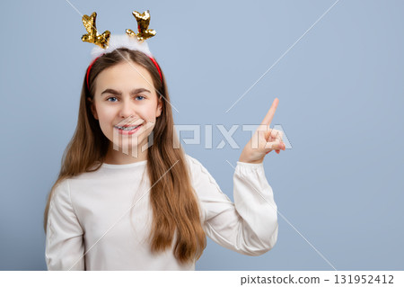 Smiling girl with sequin reindeer antlers pointing up on blue background. Festive portrait for Christmas and winter celebrations. Smiling girl with sequin reindeer antlers pointing up on blue background. Festive portrait for Christmas and winter celebrations. 131952412