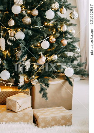 Illuminated Christmas tree with white and gold ornaments. Gifts wrapped in craft paper under evergreen branches. Festive holiday decoration for Merry Christmas. Illuminated Christmas tree with white and gold ornaments. Gifts wrapped in craft paper under evergreen branches. Festive holiday decoration for Merry Christmas. 131952987
