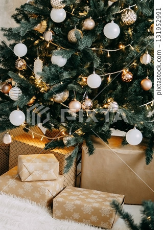 Illuminated Christmas tree with white and gold ornaments. Gifts wrapped in craft paper under evergreen branches. Festive holiday decoration for Merry Christmas. 131952991