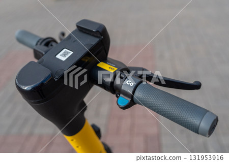 Steering wheel of an electric scooter, closeup. Rubber handlebar grips. Close up image of a steering wheel of a yellow black electric scooter. Electric scooter for rental parked on sidewalk. Steering wheel of an electric scooter, closeup. Rubber handlebar grips. Close up image of a steering wheel of a yellow black electric scooter. Electric scooter for rental parked on sidewalk. 131953916