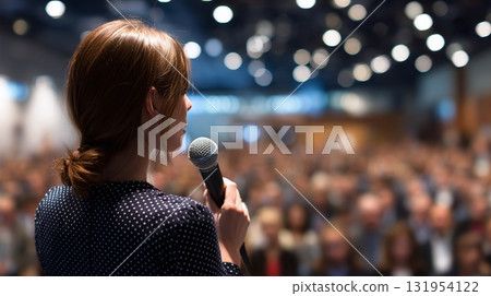 Speaker addressing crowd: A woman at a podium with a microphone captivating an audience in a large auditorium. Spotlight on public speaking and leadership. Speaker addressing crowd: A woman at a podium with a microphone captivating an audience in a large auditorium. Spotlight on public speaking and leadership. 131954122