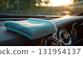 A light blue microfiber cleaning towel sits on the shiny black dashboard of a car as the sun shines through the front window in the background. 131954137