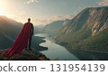 A figure in business attire and a flowing red cape stands atop a peak, gazing over a vast mountain landscape, symbolizing ambition and vision. 131954139