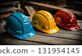 Three colorful protective hard hats in blue, yellow, and red, sitting on a weathered wooden surface with blurred wooden planks in the background. 131954145