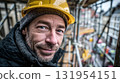 Portrait of a construction worker in a yellow hard hat, smiling slightly, with scaffolding and blurred buildings in the background. 131954151