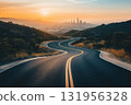 Winding road is leading through a mountain landscape toward a city skyline at sunset 131956328