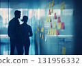 Two business colleagues are having a conversation in an office building hallway while looking at a glass wall with adhesive notes 131956332