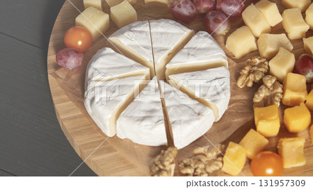A wooden board with a wedge of cheese and a variety of fruits and nuts 131957309