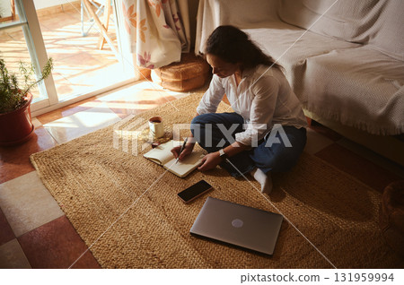 Woman Writing in Notebook at Home With Laptop, Coffee, and Sunlit Living Room Woman Writing in Notebook at Home With Laptop, Coffee, and Sunlit Living Room 131959994