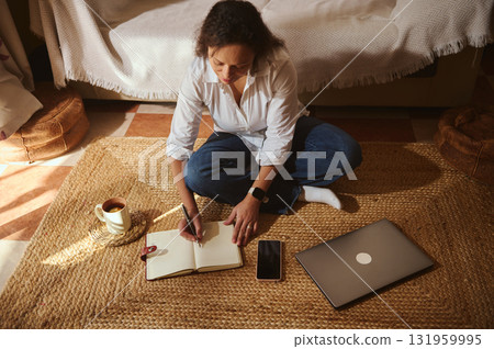 Woman Working at Home on a Rug, Writing in Notebook With Laptop and Phone Nearby Woman Working at Home on a Rug, Writing in Notebook With Laptop and Phone Nearby 131959995