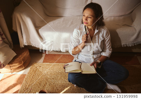 Thoughtful Woman Sits Cross-L-legged on Rug With Notebook in Sunlit Living Room Thoughtful Woman Sits Cross-L-legged on Rug With Notebook in Sunlit Living Room 131959998