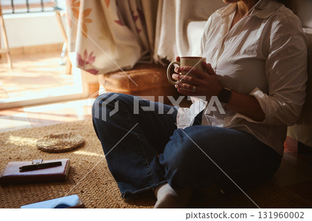 Relaxed Home Coffee Moment: Woman Sits on Floor With Mug in Sunlit Cozy Space Relaxed Home Coffee Moment: Woman Sits on Floor With Mug in Sunlit Cozy Space 131960002
