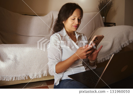 Woman Working From Home With Laptop And Smartphone, Smiling While Texting Woman Working From Home With Laptop And Smartphone, Smiling While Texting 131960050