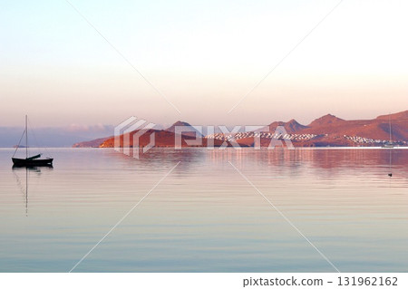 A peaceful sunrise or sunset over a calm, reflective sea, with sailboats and a small island in the distance, bathed in soft pastel light against a backdrop of mountains and a distant coastal town. A peaceful sunrise or sunset over a calm, reflective sea, with sailboats and a small island in the distance, bathed in soft pastel light against a backdrop of mountains and a distant coastal town. 131962162