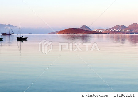A peaceful sunrise or sunset over a calm, reflective sea, with sailboats and a small island in the distance, bathed in soft pastel light against a backdrop of mountains and a distant coastal town. A peaceful sunrise or sunset over a calm, reflective sea, with sailboats and a small island in the distance, bathed in soft pastel light against a backdrop of mountains and a distant coastal town. 131962191