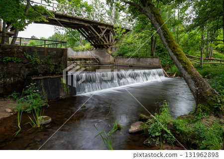 A serene and calm river gracefully flows beneath a rustic wooden bridge, enveloped by lush greenery and tranquil nature A serene and calm river gracefully flows beneath a rustic wooden bridge, enveloped by lush greenery and tranquil nature 131962898
