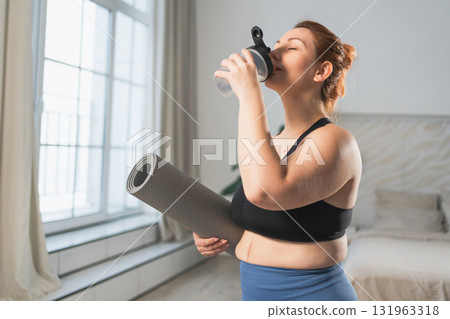 Yoga fitness sport workout training. Woman holding yoga mat drinking bottle water ready for practicing yoga fitness at home. Plus size girl in sportswear have training workout for weight loss indoor Yoga fitness sport workout training. Woman holding yoga mat drinking bottle water ready for practicing yoga fitness at home. Plus size girl in sportswear have training workout for weight loss indoor 131963318