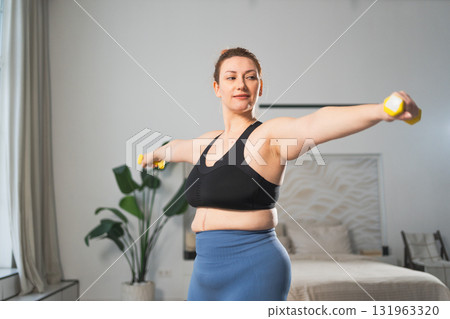 Fitness workout training. Woman picking up dumbbells. Plus size female doing sports exercise at home indoor. Girl in sportswear have workout weight training for weight loss. Sport and fitness Fitness workout training. Woman picking up dumbbells. Plus size female doing sports exercise at home indoor. Girl in sportswear have workout weight training for weight loss. Sport and fitness 131963320