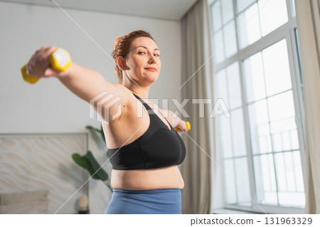 Fitness workout training. Woman picking up dumbbells. Plus size female doing sports exercise at home indoor. Girl in sportswear have workout weight training for weight loss. Sport and fitness 131963329