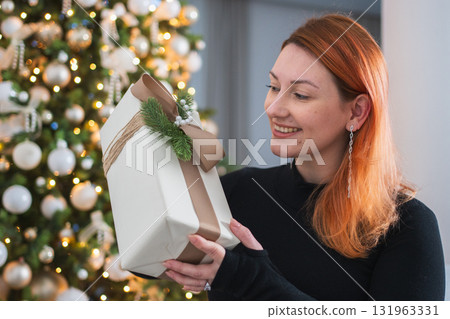 Merry Christmas. Woman unpacking open up gift box near Christmas tree. Girl in living room with Christmas tree holding opening gift box with surprise face. Woman celebrating Christmas eve at home 131963331