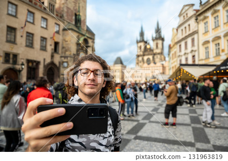 Prague, Czech Republic, August 7, 2023. A young Caucasian man takes a selfie with his cell phone in the iconic and crowded Old Town Square. Using technology. Blurred background, travel destinations. 131963819
