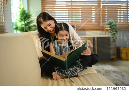 An Asian mother and daughter sit together indoors, smiling and bonding over a book 131963983