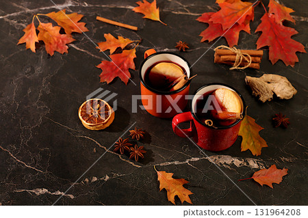 Two colorful enamel mugs with hot autumn drink, mulled wine with apples and autumn fallen leaves, star anise and cinnamon on a marble table,banner for display,autumn greeting concept, mockup for menu 131964208