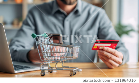 Man using a laptop and smartphone to make an online payment, holding a red credit card. A miniature shopping cart symbolizes e-commerce and digital transaction concepts 131964310