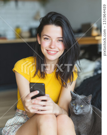 Smiling young woman with dark hair in yellow t-shirt and plaid shorts sitting on the floor, holding a smartphone next to her grey cat, capturing casual, relaxed moment of domestic life, pet ownership 131964489