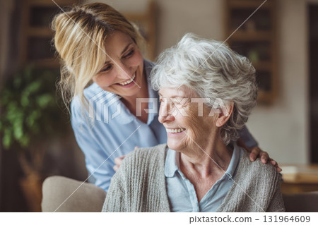 A warm, candid moment showing an elderly woman and a middle-aged woman sharing a smile and affectionate bond. The younger woman stands behind the senior, placing her hands on her shoulders 131964609