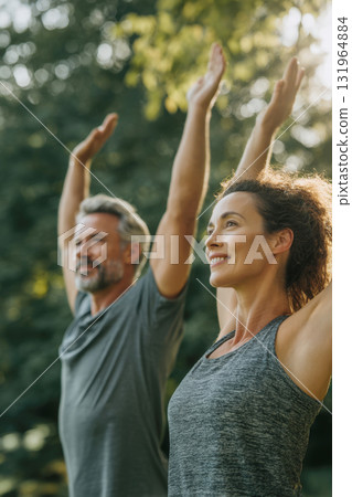 Mature couple practices yoga together in park on sunny day. With arms raised, serene smile, focus is on the woman in the foreground. Themes of essence of a healthy, active lifestyle, outdoor fitness Mature couple practices yoga together in park on sunny day. With arms raised, serene smile, focus is on the woman in the foreground. Themes of essence of a healthy, active lifestyle, outdoor fitness 131964884