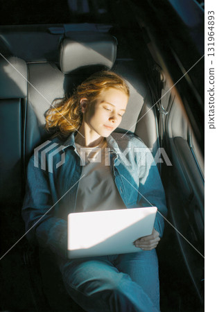 young woman with red hair sits in backseat of car, using her laptop in moment of focused work or creativity. Sunlight streams through window, device, suggesting a modern approach to work from anywhere 131964893