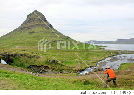 A photographer in an orange jacket captures the iconic Kirkjufell mountain in Iceland, A photographer in an orange jacket captures the iconic Kirkjufell mountain in Iceland, 131964980