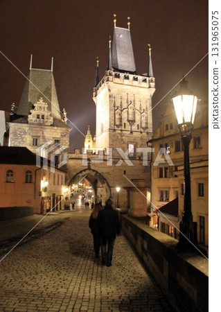 Nighttime scene on Prague historic Charles Bridge with a couple walking toward the illuminated Old Town Bridge Tower, surrounded by gothic architecture and glowing streetlamps 131965075