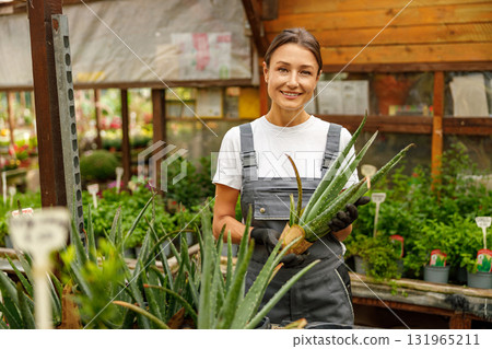A Smiling Woman in a Greenhouse Surrounded by Lush Aloe Vera Plants and Vibrant Flora 131965211