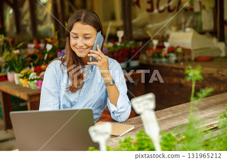 A young woman works on her laptop while talking on the phone in a vibrant flower shop A young woman works on her laptop while talking on the phone in a vibrant flower shop 131965212