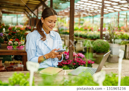 A professional woman is working diligently in a vibrant flower shop using her laptop and phone 131965236