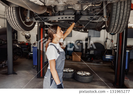 A skilled woman mechanic is carefully inspecting a vehicle in an automotive workshop environment A skilled woman mechanic is carefully inspecting a vehicle in an automotive workshop environment 131965277