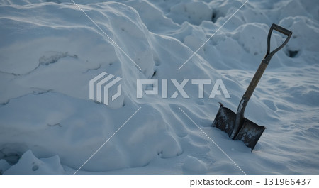 An old metal shovel with a wooden handle is stuck upright in a large, textured pile of fresh white snow under cold winter light, suggesting hard work and seasonal conditions. An old metal shovel with a wooden handle is stuck upright in a large, textured pile of fresh white snow under cold winter light, suggesting hard work and seasonal conditions. 131966437