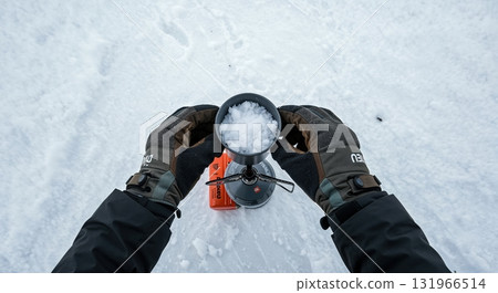 A person's dark gloved hands hold a silver pot full of white snow over a small orange camping stove, preparing water in a vast snowy outdoor environment. 131966514