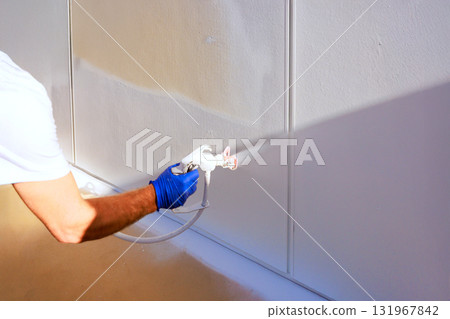Handyman is applying fresh white paint to wall using spray gun in well lit room. Handyman is applying fresh white paint to wall using spray gun in well lit room. 131967842