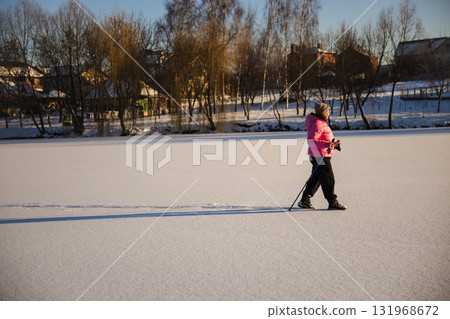 Elderly woman walking with trekking poles on a snowy frozen field in bright winter sunlight. She stays active and healthy, enjoying exercise and fresh air during a peaceful day outdoors. Elderly woman walking with trekking poles on a snowy frozen field in bright winter sunlight. She stays active and healthy, enjoying exercise and fresh air during a peaceful day outdoors. 131968672