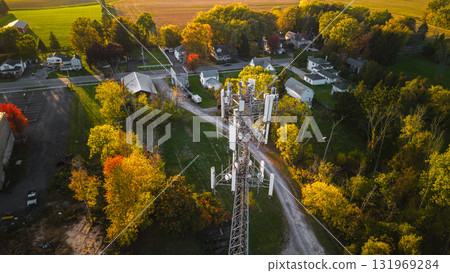 Communication tower near small suburban houses surrounded by autumn trees and green fields, captured from above in warm evening light. Communication tower near small suburban houses surrounded by autumn trees and green fields, captured from above in warm evening light. 131969284