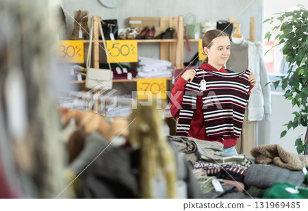 European happy teen girl buyer standing in clothing market and carefully examines selection of sweaters on Black Friday sale of winter clothing collection European happy teen girl buyer standing in clothing market and carefully examines selection of sweaters on Black Friday sale of winter clothing collection 131969485