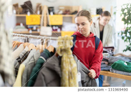 Positive young girl standing in interior of clothing boutique and choosing something on Black Friday sale of winter clothing collection 131969490