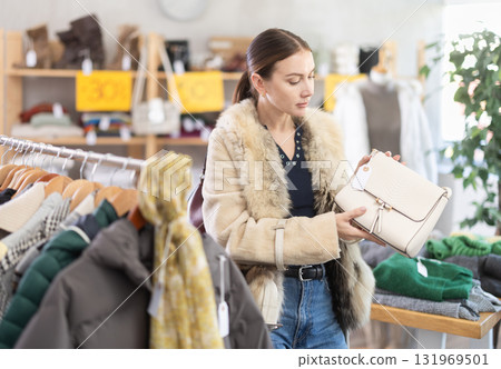 Black Friday sale of winter clothing collection. Positive woman customer standing against background of shelves with clothes in modern store and choosing bag Black Friday sale of winter clothing collection. Positive woman customer standing against background of shelves with clothes in modern store and choosing bag 131969501